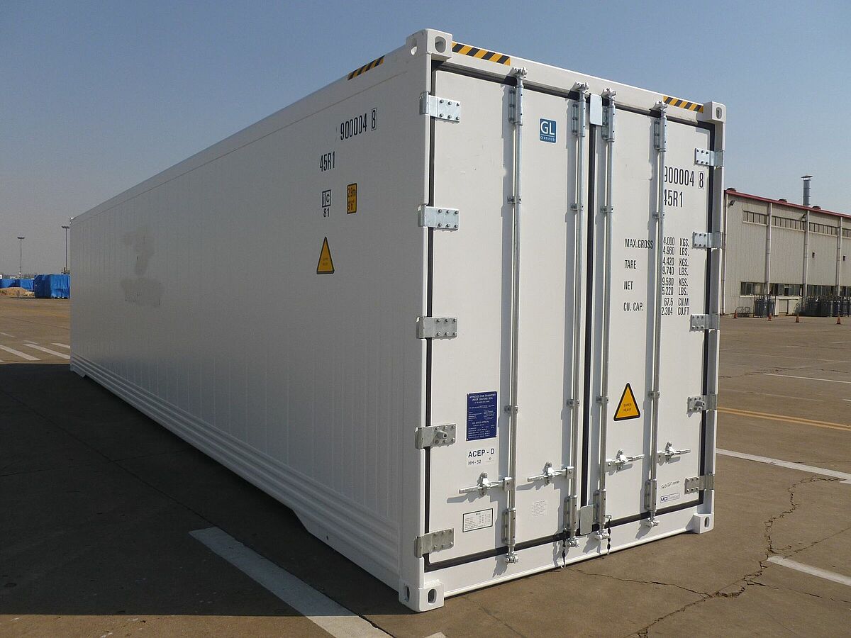 Ein weißer 40-Fuß-Kühlcontainer steht auf einem weitläufigen Industriegelände mit Fahrbahnmarkierungen. Die Containeraußenseite ist glatt, mit verstärkten Ecken, Verriegelungsstangen und mehreren Sicherheits- und Informationskennzeichnungen, darunter Warnsymbole und technische Angaben zu Gewicht und Volumen. Im Hintergrund sind Lagerhallen und einige abgedeckte Objekte sichtbar.