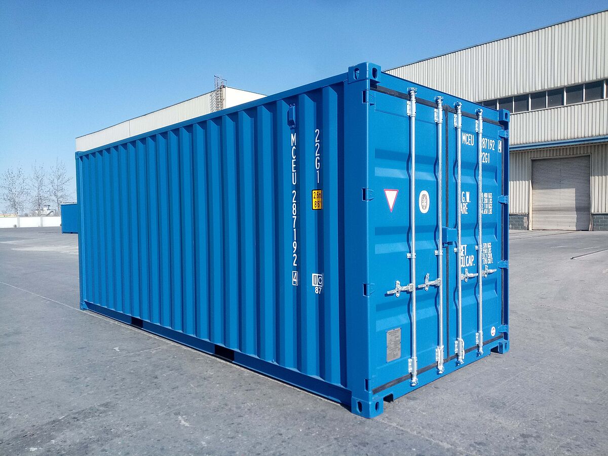 Ein blauer 20-Fuß-Standard-Seecontainer steht auf einer asphaltierten Freifläche vor einer Industriehalle. Der Container besitzt verstärkte Ecken, Verriegelungsstangen an der Tür und ist mit verschiedenen technischen Kennzeichnungen und Nummern beschriftet.